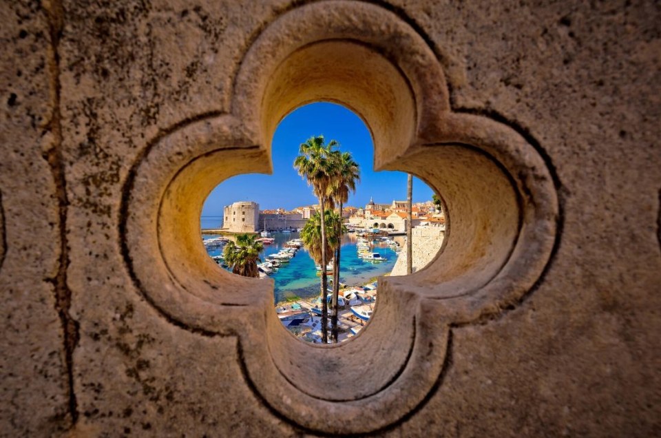 View of a coastal city through a stone window with palm trees and boats.