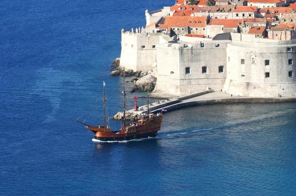A wooden sailboat near fortified city walls by the sea.