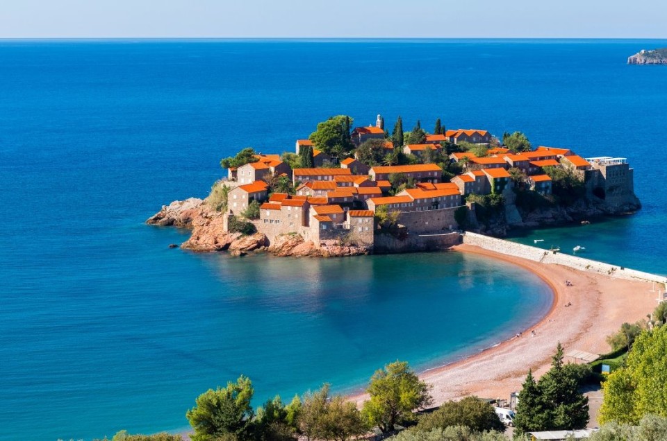Island with red-roofed buildings surrounded by blue sea and sandy beach.